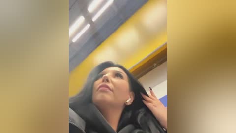 Media: A candid video of a woman with dark hair and fair skin, wearing a black jacket, looking up thoughtfully. Background includes yellow and white walls, fluorescent lights, and blurred figures.