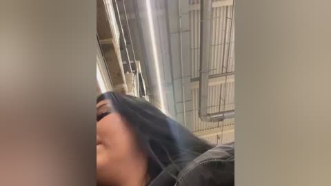 Media: Video of a woman with medium skin tone and dark hair, wearing a gray jacket, lying on a hospital bed with a metallic railing and white sheets, in a dimly lit room.