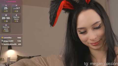 Media: Video of a young woman with long black hair, wearing a red and black bunny ear headband, smiling. Background includes a beige wall and a soft, blurred couch.