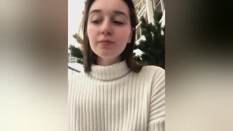 Media: Video of a young, fair-skinned woman with straight, shoulder-length brown hair, wearing a white, ribbed-knit turtleneck sweater, standing indoors with a Christmas tree in the background.