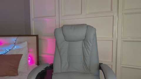 Media: Video of a modern, gray leather office chair with a padded backrest in a minimalist room with white paneled walls, a pink and blue LED strip light, and a white bed with a brown pillow.