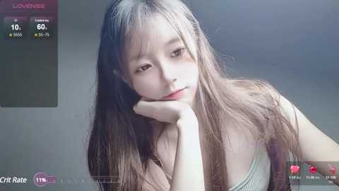 Media: Video of a young Asian woman with long, straight brown hair, fair skin, and a slender figure. She wears a light-colored top and leans on her elbow, looking contemplative against a plain background.