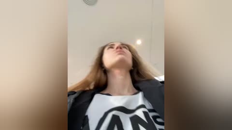 Media: A video of a young woman with long, straight, light brown hair, wearing a black jacket over a white shirt with bold black text, gazing upward in a minimalist, beige-walled room with a ceiling light.