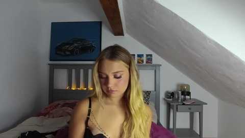 Media: A video of a young blonde woman with long hair, wearing a black and beige bra, sitting on a bed in a cozy attic room.