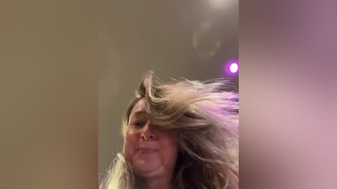 Media: Video of a woman with long, blonde hair, blowing it upwards, creating a dynamic motion blur. The background is a blurred, beige wall with a purple light source in the top right corner.