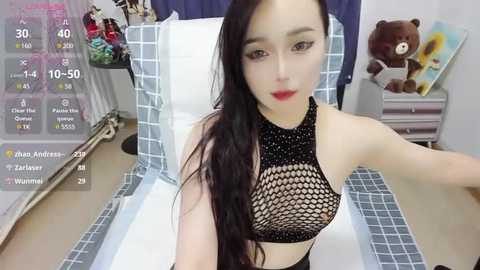 Media: A video of an Asian woman with long black hair, wearing a black fishnet top, in a bedroom with a white bed and teddy bear.