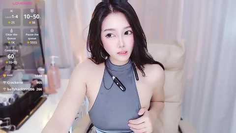 Media: Video of a young East Asian woman with long black hair, wearing a grey halter top, sitting in a modern, well-lit room.