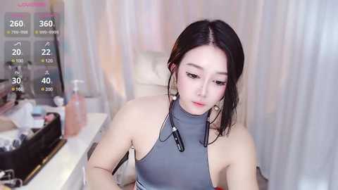 Media: Video of an East Asian woman with fair skin, wearing a gray halter top, sitting in a modern room with a white couch and pink decor.