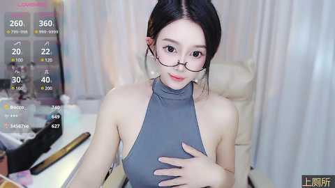 Media: Video of a fair-skinned Asian woman with long black hair in a bun, wearing glasses, a grey halter top, and a white pearl necklace, seated at a white desk in a well-lit room.