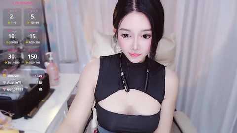 Media: Video of an East Asian woman with pale skin, black hair, and a black, cut-out top, sitting on a white chair, with a digital screen displaying metrics in the background.