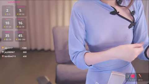 Media: Video of a woman in a light blue blouse, wearing a headset, in a living room with beige curtains and a gray couch.