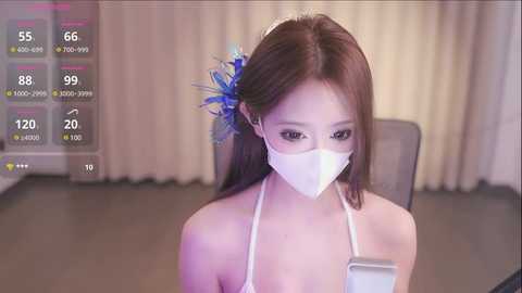 Media: A video of an Asian woman with fair skin and long brown hair, wearing a white face mask and a blue flower accessory. She's seated indoors, with a digital overlay showing a virtual temperature of 66\u00b0F.