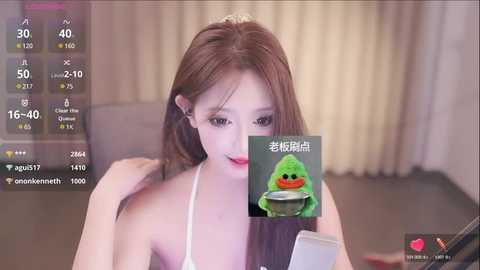 Media: A young Asian woman with long brown hair, wearing a white top, sits at a desk, holding a phone. The background shows a blurry room with beige curtains. A digital overlay displays a green frog and Chinese text.