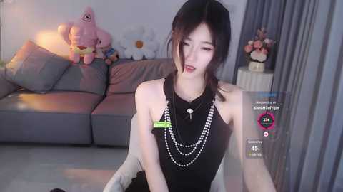 Media: Video of an Asian woman with fair skin, wearing a black sleeveless top, multiple necklaces, and a green hair tie, seated on a gray couch in a modern living room.