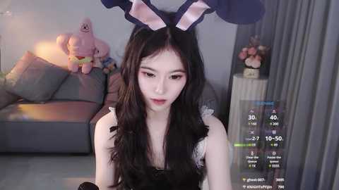 Media: Video of a young East Asian woman with long black hair, wearing bunny ears and a black lace dress, in a cozy, dimly lit living room with a gray couch and plush toys.