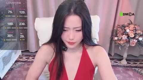 Media: Video of an East Asian woman with long black hair, wearing a red halter dress, seated on a couch. Background includes floral arrangements, a window, and digital camera display.