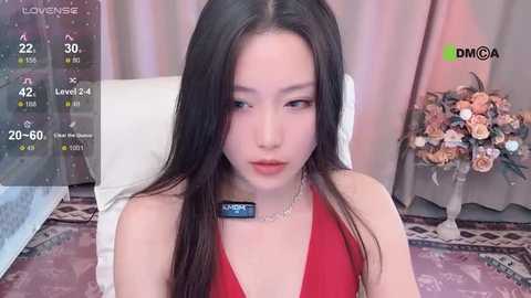 Media: Video of a young East Asian woman with long black hair, wearing a red halter top, sitting on a bed with a white headboard, against a pink curtain background.