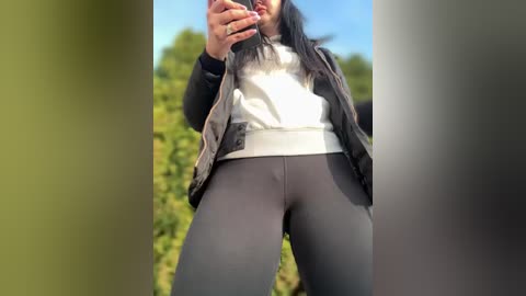 Media: Video of a woman in a white top and black jacket, holding a phone, standing in front of greenery.