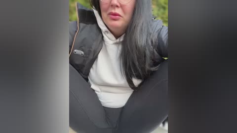 Media: A video of a woman with long black hair and glasses, wearing a black puffer jacket over a white turtleneck, crouching with legs apart, partially obscured by dark, vertical bars.