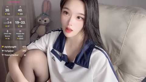 Media: Video of an East Asian woman with long black hair, fair skin, and small breasts, wearing a white polo shirt with blue accents, sitting on a beige couch. Background features a stuffed bunny.