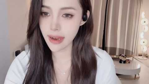 Media: Video of a young woman with pale skin, long dark hair, and a slender build, wearing a white top and black headphones. She has a neutral expression. Background shows a modern room with beige curtains and a mirror with lights.