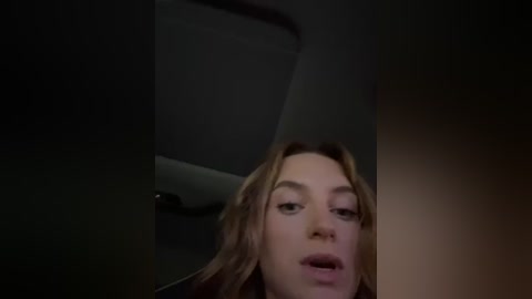 Media: Video of a woman with light skin and long brown hair, looking slightly up, her mouth slightly open, taken from a low angle in a dimly lit room.