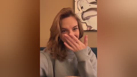 Media: Video of a smiling woman with shoulder-length, wavy brown hair, covering her mouth with her right hand. She wears a light gray sweatshirt. Background features a beige wall with a modern abstract painting.