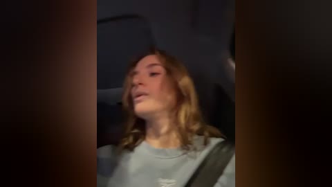 Media: Video of a young woman with shoulder-length blonde hair, wearing a light gray shirt, seated in a dimly lit car, with blurred background.