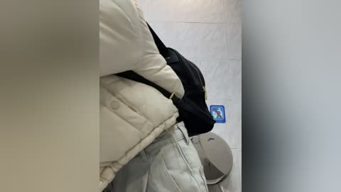 Media: Video of a person in a beige quilted jacket and light blue jeans sitting on a grey tiled floor in a bathroom. Black backpack on the left.