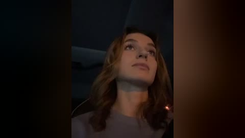 Media: Video of a young woman with light skin and wavy brown hair, wearing a grey top, looking upward with a contemplative expression, dimly lit interior background.