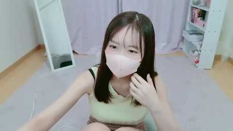 Media: Video of an East Asian girl with long black hair, wearing a light green sleeveless top and a pink face mask, seated on a light carpet in a room with white walls and a purple curtain backdrop.