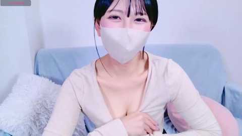 Media: Video of an East Asian woman with short black hair, wearing a beige cardigan, white face mask, and sitting on a light blue couch.