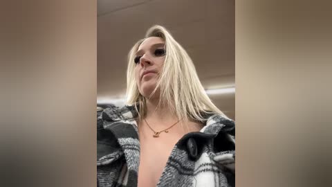 Media: Video of a blonde woman with a fair complexion, wearing a black and white plaid shirt, standing indoors, looking down, with blurred background.