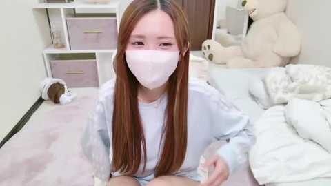 Media: A video of a young Asian woman with long, straight brown hair, wearing a white face mask and a light blue sweater, sitting on a bed in a soft, pastel-colored bedroom with stuffed animals.