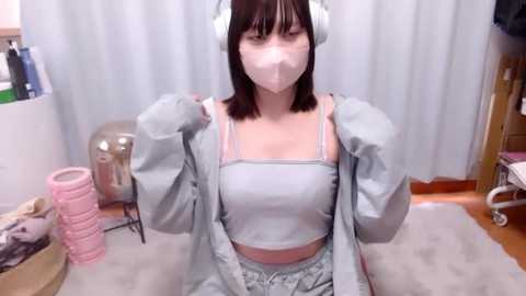 Media: Video of a young Asian woman with short black hair, wearing white headphones, a light blue crop top, and matching pajama pants, seated indoors on a fluffy rug, with a white curtain backdrop and various household items in the background.