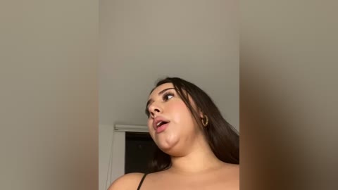 Media: Video of a plus-size woman with long dark hair, wearing a black top, looking up in surprise, against a plain beige wall with a window.