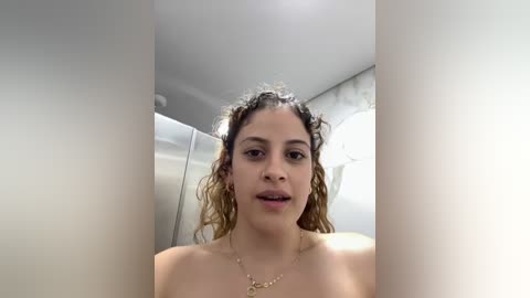 Media: Video of a topless, curly-haired, light-skinned woman with light makeup, wearing a gold necklace, in a modern bathroom with white marble tiles and a round, white light fixture.