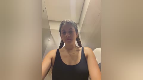 Media: Video of a young woman with medium brown skin and braided hair, wearing a black tank top, standing in a modern, minimalist bathroom with beige walls and recessed lighting.