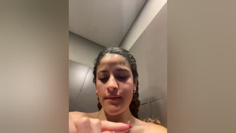 Media: Video of a young woman with light brown skin, curly dark hair, and closed eyes, standing in a tiled shower with beige walls, focusing on a droplet of water on her finger.