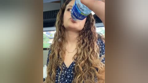 Media: Video of a young woman with long, wavy, light brown hair, wearing a blue floral dress, drinking from a blue Poland Spring bottle, blurred background with a TV displaying greenery.