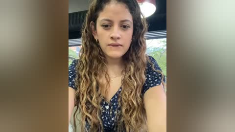 Media: A video of a young Latina woman with long, wavy, light brown hair, wearing a navy blue floral top, standing indoors with a blurred background featuring a window and greenery.