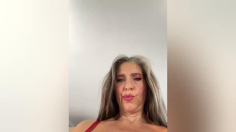 Media: Video of a middle-aged woman with long, straight gray hair and bright red lipstick, wearing a red bra, against a white background.