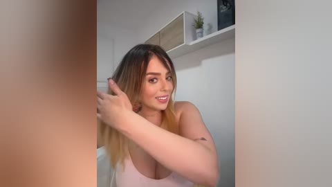 Media: Video of a young woman with fair skin and long, straight, blonde hair, styled with a hair dryer. She has a slim physique and wears a light pink tank top. The background shows a white wall with a shelf and a small potted plant.