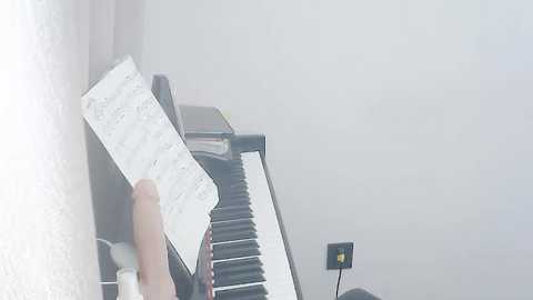 Media: A close-up video of a hand holding a sheet of music, with a piano and a light switch in the background, set against a plain, light-colored wall.