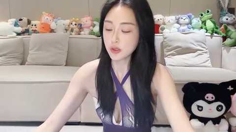 Media: Video of an Asian woman with long black hair, wearing a purple halter top, sitting on a beige couch. Background includes stuffed animals and a plush bear.