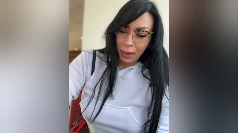 Media: Video of a young woman with long black hair, wearing glasses and a white hoodie, looking down, possibly in a bathroom, with blurred faces in the background.