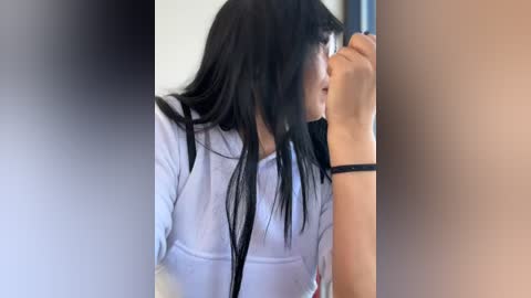 Media: Video of a young woman with long black hair, wearing a white shirt, standing in a blurred, beige-colored room, holding a black bracelet.