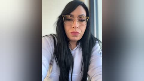 Media: Video of a young woman with long, straight black hair, wearing large, rectangular glasses and a light-colored top, looking serious and focused.