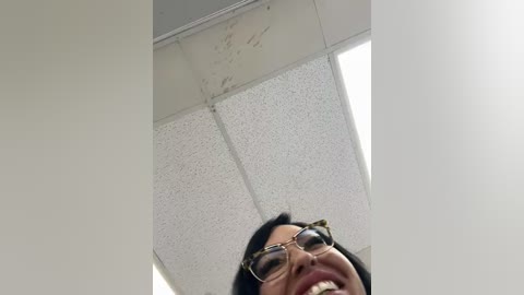 Media: Video of a smiling Asian woman with glasses, standing in a fluorescent-lit office with a white ceiling and grid pattern, looking upwards.