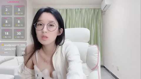 Media: Video of an East Asian woman with long black hair, wearing glasses and a white cardigan, seated in a modern, white room with a white chair and green curtains.